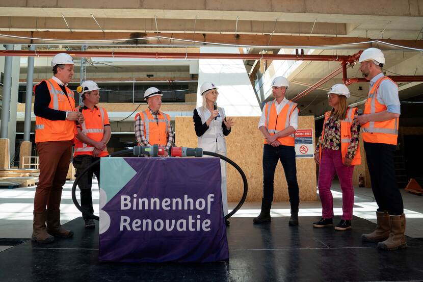Mona Keijzer met medewerkers van de aannemers en het Rijksvastgoedbedrijf op het podium in de Statenpassage van de Tweede Kamer