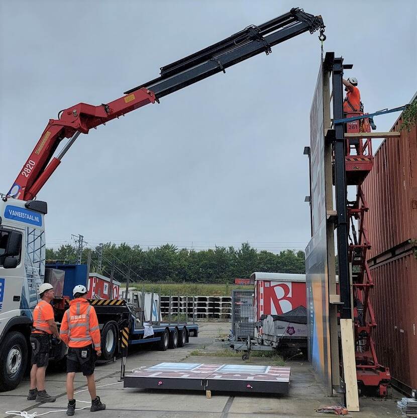 Inhijsen van een paneel door een mobiele bouwkraan, met rechts achter de bouwschutting een medewerker op een hoogwerker